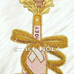 May include: An embroidered patch featuring a pink champagne bottle with a gold glitter bow, a cork popping, and gold glitter spray. The label reads MOET CHANDON with a heart and star symbol. The text STITCH NOLA is visible at the top and center.
