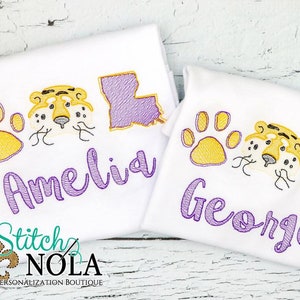 May include: Two white t-shirts with purple embroidery. The shirts feature a paw print, a tiger face, and the state of Louisiana. The shirts are personalized with the names "Amelia" and "George".