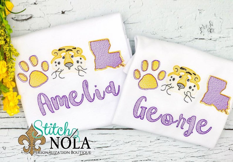 May include: Two white t-shirts with purple embroidery. The shirts feature a paw print, a tiger face, and the state of Louisiana. The shirts are personalized with the names "Amelia" and "George".