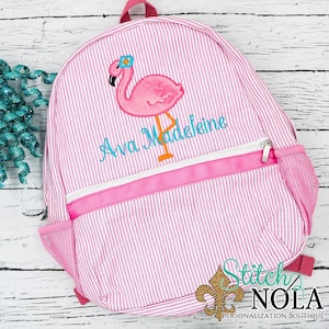 May include: Pink and white striped backpack with a pink flamingo embroidered on the front. The backpack has a pink zipper and a mesh pocket on the side. The text "Ava Madeleine" is embroidered below the flamingo.