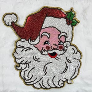 May include: A decorative patch featuring a beaded Santa Claus face with a red hat, white beard, and mustache. The design includes a green holly leaf with red berries on the hat, all outlined in gold trim, crafted with small, shiny sequins.