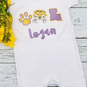 May include: White baby onesie with purple and gold embroidery of a paw print, tiger head, and the state of Louisiana. The name "logan" is embroidered in purple below the design.