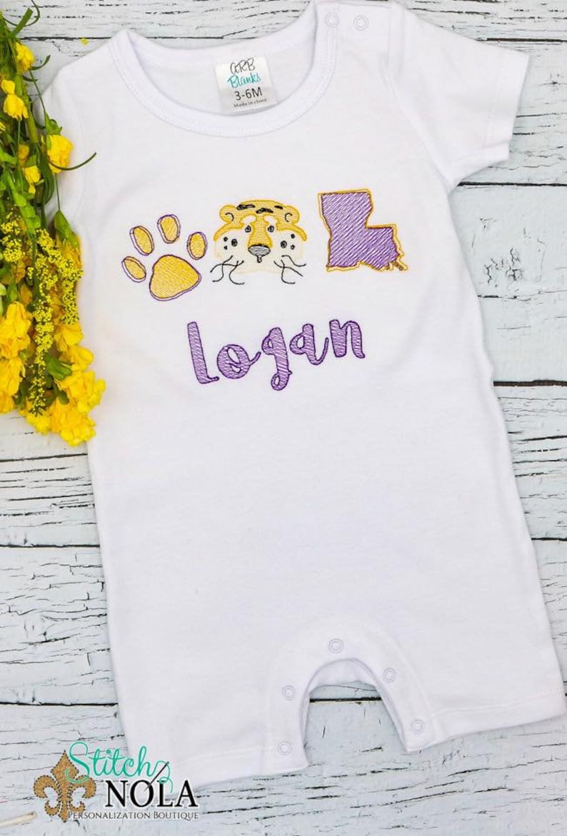 May include: White baby onesie with purple and gold embroidery of a paw print, tiger head, and the state of Louisiana. The name "logan" is embroidered in purple below the design.