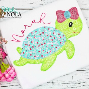 May include: A white t-shirt with a green and blue embroidered turtle with a pink bow. The name "Noah" is embroidered in pink above the turtle.