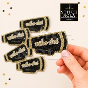 May include: A collection of black and gold glitter megaphone-shaped patches featuring the text who dat in a cursive font. The patches have a textured, beaded appearance with gold glitter borders and black centers, arranged in a scattered pattern.