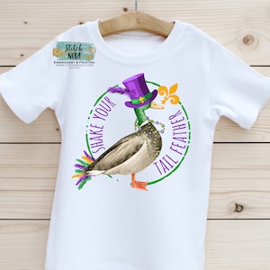 May include: White t-shirt with a green, purple, and gold illustration of a duck wearing a purple hat and beads. The text "Shake Your Tail Feather" is written around the duck.