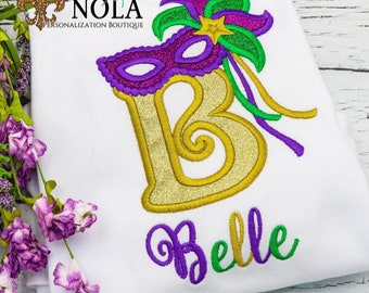 May include: A gold glitter letter B with a purple and green Mardi Gras mask and feather decoration. The name "Belle" is embroidered below in purple and green.