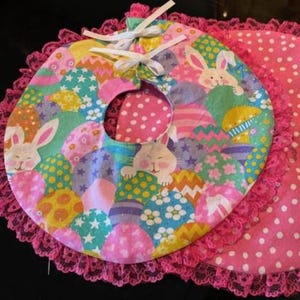 May include: A pink and white fabric bib with a pink lace trim. The bib features a colorful Easter egg pattern with bunnies and stars.