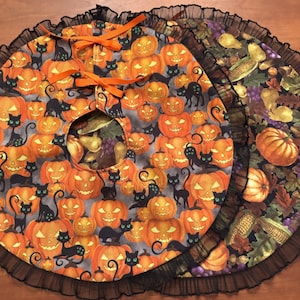 May include: A circular fabric tree skirt featuring a Halloween print of orange jack-o'-lanterns and black cats on a gray background, finished with a black ruffled edge and orange ribbon ties. A second skirt with a harvest print is layered underneath.