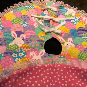 Op de afbeelding: Een roze en witte kerstboomrok met een kantversiering en een gat in het midden voor de boomstam. De rok is versierd met kleurrijke paaseieren en konijnen.