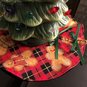 May include: A red and black plaid Christmas tree skirt with gingerbread men designs. The skirt has a green ribbon tied around it.