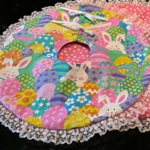 May include: A pink and white fabric Easter tree skirt with a lace trim. The skirt features a pattern of colorful Easter eggs and bunnies.