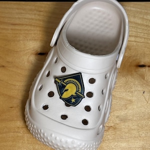 May include: A white clog with a gold and blue pin featuring a Spartan helmet and a sword. The pin is on the front of the clog.