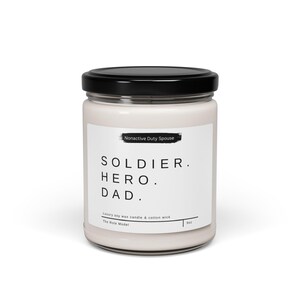 Può includere: Un barattolo di candela bianco con un coperchio nero. L'etichetta recita "Nonactive Duty Spouse" e "SOLDIER. HERO. DAD." con il testo "Luxury soy wax candle & cotton wick" e "The Role Model" sotto.