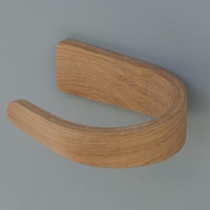 May include: A wooden, curved toilet paper holder. The holder is made of light-colored wood and has a smooth, curved design. It is mounted on a gray wall.