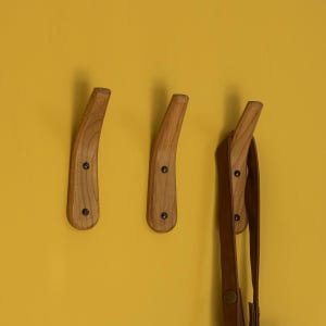 May include: Three wooden wall hooks with a curved design, mounted on a yellow wall. A brown leather strap hangs from one of the hooks.