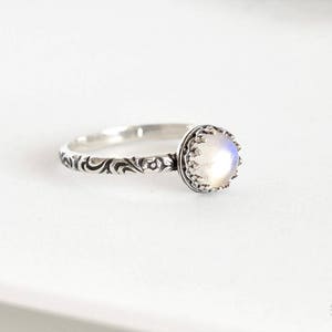 May include: A silver ring with a delicate floral design and a white moonstone set in a crown-like setting.