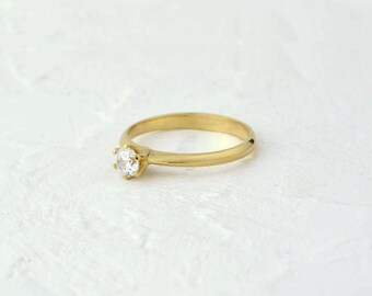 May include: A gold band ring with a single, clear, round gemstone.