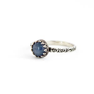 May include: A silver ring with a blue gemstone set in a crown-like setting. The band is decorated with a swirling pattern.