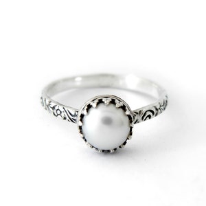 May include: A silver ring with a white pearl set in a decorative bezel. The band is adorned with an intricate floral design.