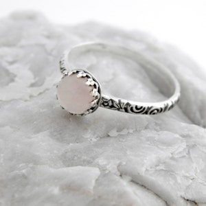 May include: A delicate silver ring with a pink gemstone set in a crown-like setting. The band is adorned with a floral design.