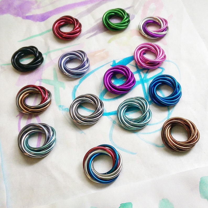 May include: A collection of 13 colorful metal rings, each with a unique design. The rings are arranged on a white surface with a light purple and blue watercolor background.
