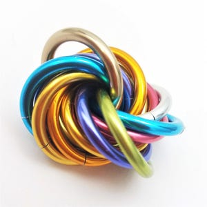 May include: A colorful metal puzzle featuring interlocking rings in various shades, including gold, blue, purple, pink, and silver. The rings are arranged in a spherical shape, creating a visually appealing and intricate design.