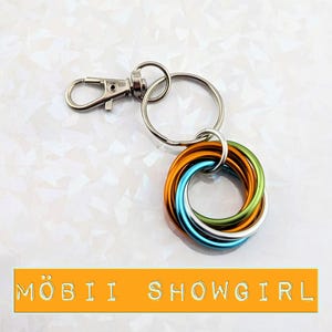 May include: A silver-colored keychain with a lobster clasp and a split ring. The keychain features a cluster of interlocking rings in orange, blue, silver, and green. The text "MÖBII SHOWGIRL" is displayed on an orange banner.