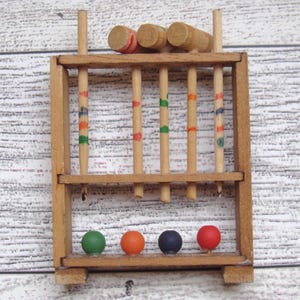 May include: A miniature wooden croquet set with a rectangular frame, vertical posts, and four colored balls: green, orange, navy, and red. The set includes wooden mallets and striped posts. The background is a light wood grain.