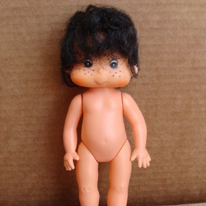May include: A vintage doll with a peach-colored body, black hair, and freckles. The doll is standing upright with arms at its sides. The doll is approximately 5 inches tall.