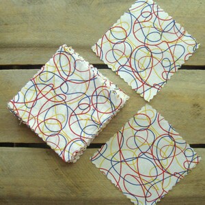 May include: Three white square coasters with a colorful abstract design of red, blue, and yellow lines. Each coaster has a scalloped edge. The coasters are arranged on a wooden surface.