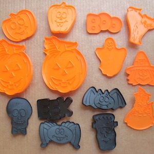 May include: A collection of thirteen plastic Halloween cookie cutters in orange and black. The shapes include pumpkins, a ghost, a cat, a witch, a skull, a bat, a spider, Frankenstein's monster, and the words BOO and EEK in block lettering.