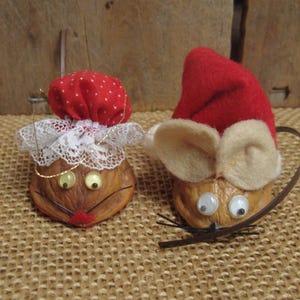 May include: Two decorative ornaments crafted from walnut shells. One mouse figure wears a red polka-dot bonnet with white lace trim. The other mouse figure wears a red felt hat and has large beige felt ears, googly eyes, and thin black whiskers.