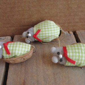 May include: Three small mouse ornaments, each crafted with a grey fuzzy body and a white bead nose, tucked into a brown walnut shell. Each mouse is wrapped in a light green and white gingham fabric blanket secured with a thin red ribbon.