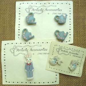 May include: A collection of decorative buttons on three cards labeled Artistic Accessories. The set includes four light blue watering cans with pink floral accents, one standing rabbit in a blue suit with a red bowtie, and two white rabbits.