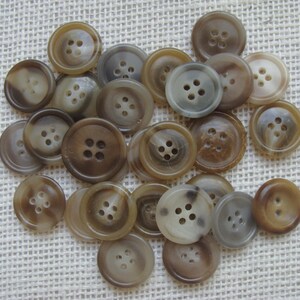 May include: A collection of 20 brown and white mottled buttons with four holes each. The buttons are arranged in a random pattern on a beige fabric background.