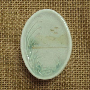 May include: An oval ceramic dish with a glossy off-white finish, featuring a hand-painted landscape design in soft green and brown tones. The illustration depicts tall grasses on the left side and distant rolling hills under a pale sky.