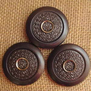 May include: Three round, dark brown wooden coasters with a raised, leaf-shaped design and a gold metal ring in the center.