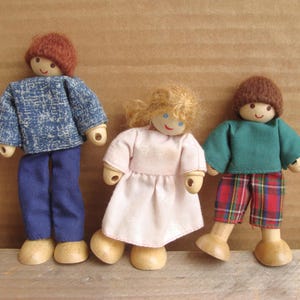 May include: Three wooden doll figures with painted faces and yarn hair. One doll wears a blue denim-look shirt and pants, another a pink dress, and the third a green shirt and plaid shorts. The dolls have wooden bodies and feet.