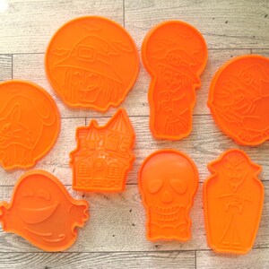 May include: A set of eight bright orange plastic cookie cutters featuring various Halloween-themed shapes including a cat, a witch, a haunted house, a ghost, a skull, and a vampire in a coffin, all arranged on a light-colored wood grain surface.