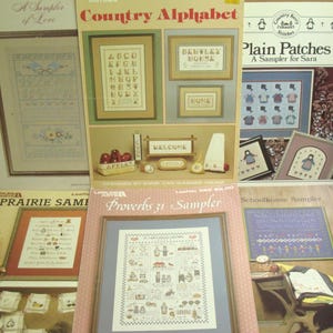 May include: A collection of six cross-stitch pattern booklets featuring titles such as A Sampler of Love, Country Alphabet, Plain Patches, Prairie Sampler, Proverbs 31 Sampler, and Schoolhouse Sampler, displaying various embroidery designs.