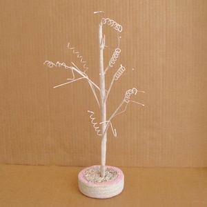 May include: A decorative white tree with wire spiral accents. The tree trunk is set in a round base with a pink and white trim. The tree is set against a brown background.