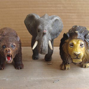 May include: Three plastic animal figurines: a brown bear with an open mouth, a gray elephant with tusks, and a lion with a yellow and brown mane. The toys are on a light brown surface.
