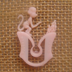 May include: A light pink plastic toy featuring a monkey figure atop a curved base with the word "Squeeze" and "Sides" printed on it. The toy has a playful design and is likely a vintage item.