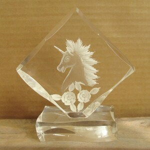May include: A clear glass sculpture of a unicorn with a detailed mane and horn, set within a diamond-shaped frame. Below the unicorn are etched roses. The sculpture sits on a rectangular base.
