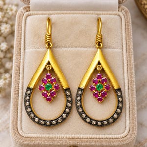 May include: A pair of gold drop earrings featuring a teardrop-shaped frame with a dark, oxidized silver border set with small clear stones. A central cluster of vibrant pink rubies surrounds a single green emerald in each earring.