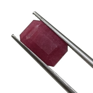 May include: A rectangular, deep pinkish-red gemstone with a faceted cut, held securely by a pair of silver-toned metal tweezers. The stone displays a semi-opaque clarity with visible natural internal inclusions and a polished surface finish.