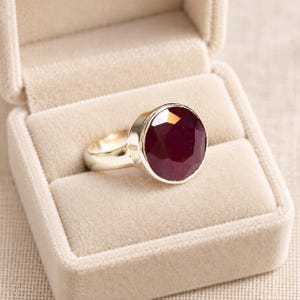 May include: A ring featuring a large, round, faceted deep red gemstone set in a polished silver-toned metal band. The ring is displayed resting inside a beige velvet-lined jewelry box, highlighting the contrast between the dark stone and metal.