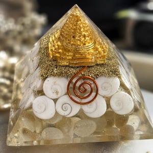 Puede incluir: Una pirámide de resina transparente que contiene un símbolo Sri Yantra dorado en la parte superior, incrustada con conchas espirales blancas, purpurina dorada y una bobina de alambre de color cobre. La base está llena de piedras claras y lisas.