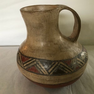 Puede incluir: Una jarra de cerámica marrón con un diseño geométrico en negro, rojo y blanco. La jarra tiene una base ancha y un cuello estrecho con una asa.